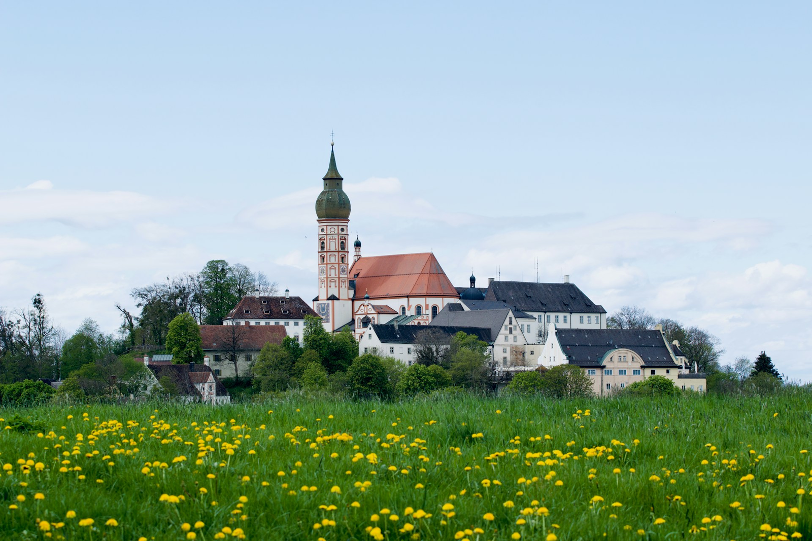 Kloster Andechs mit dem Zwiebelturm der Wallfahrtskirche auf dem Heiligen Berg, davor eine Wiese mit blühendem Löwenzahn