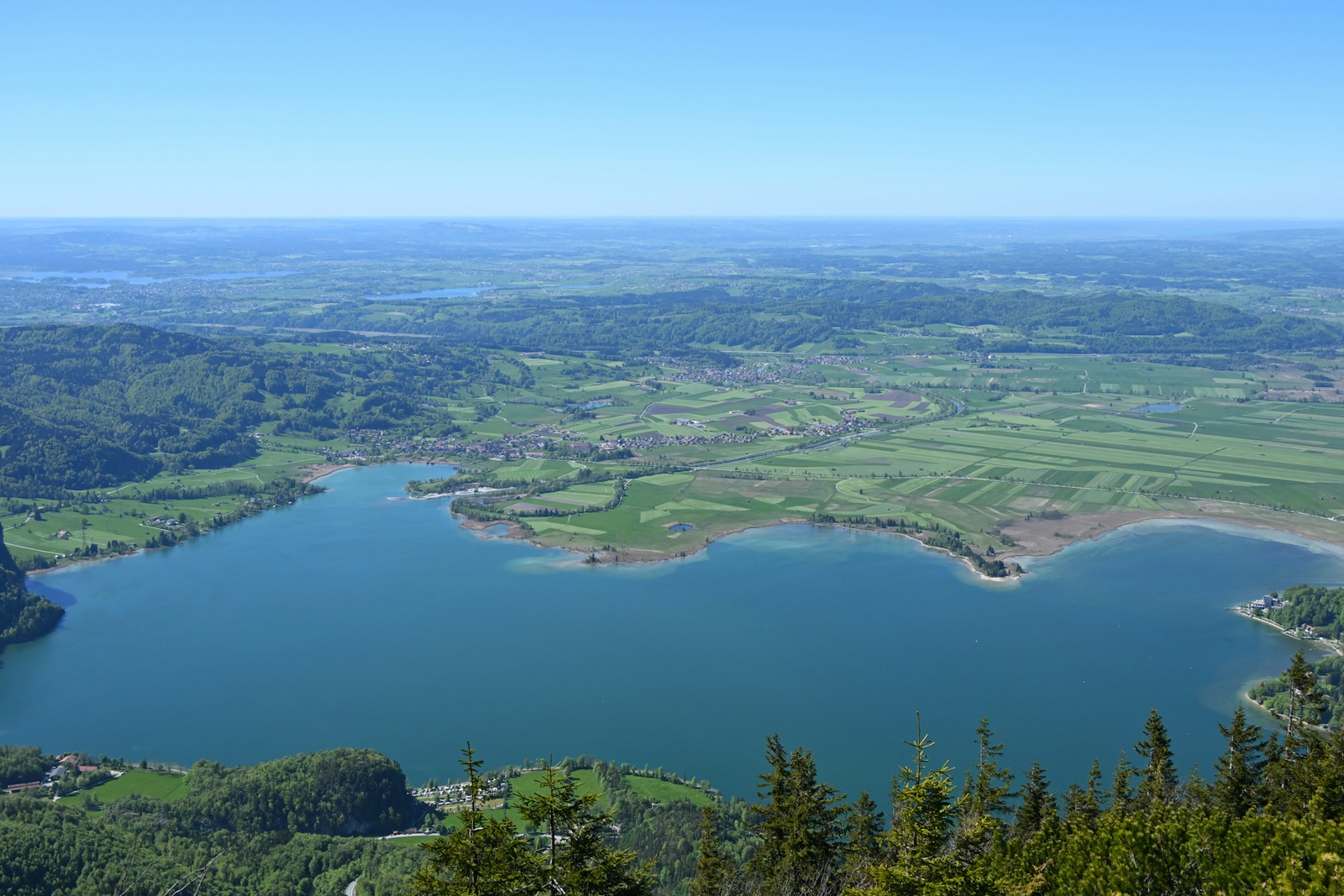 Tiefblick vom Jochberg auf den Kochelsee und die flachen grünen Wiesen des Loisach-Kochelsee-Moors, dahinter das hügelige Voralpenland