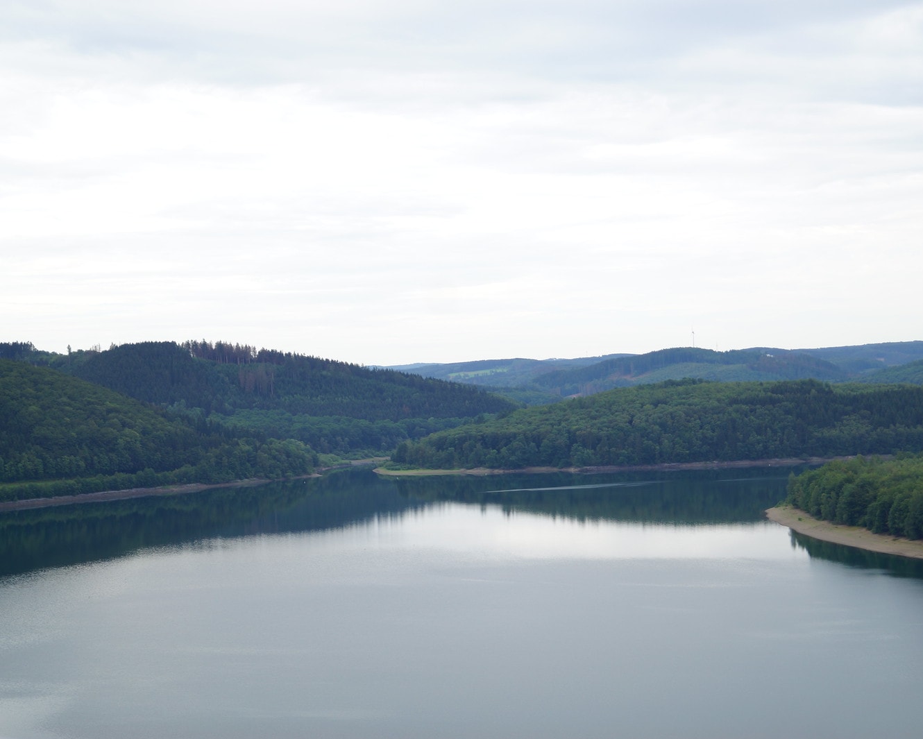 Ruhige Wasserfläche des Biggesees zwischen bewaldeten Hügeln, am rechten Ufer ein heller Uferstreifen, darüber bedeckter Himmel