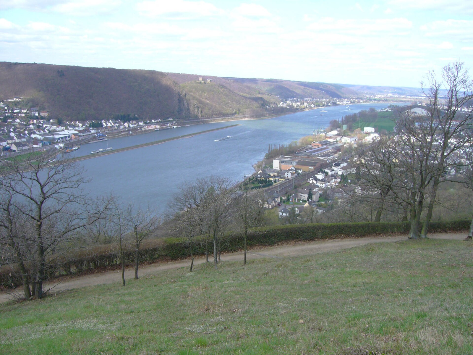 Blick von einer Wiese mit kahlen Bäumen hinab auf den Rhein; rechts die Häuser von Rheinbrohl, gegenüber ein Ort mit Hafen unter bewaldeten Hängen