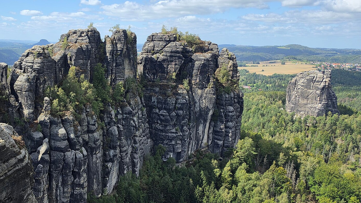 Zerklüftete Sandsteintürme der Schrammsteine über dichtem Wald, rechts der einzeln stehende Falkenstein, am Horizont Felder, ein Dorf und der Lilienstein