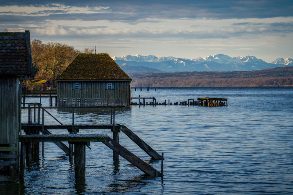 Hölzernes Bootshaus auf Pfählen im Ammersee mit Steg und weiteren Bootshäusern, am Horizont eine schneebedeckte Alpenkette
