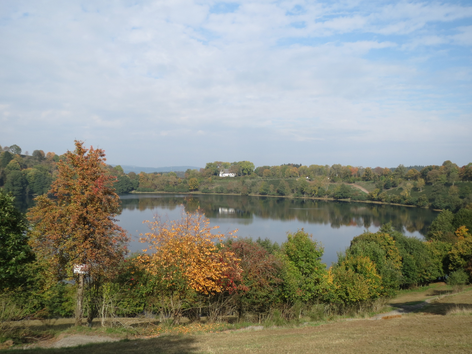 Das Weinfelder Maar im Herbst, ein runder Kratersee mit der weißen Weinfelder Kapelle am gegenüberliegenden Rand