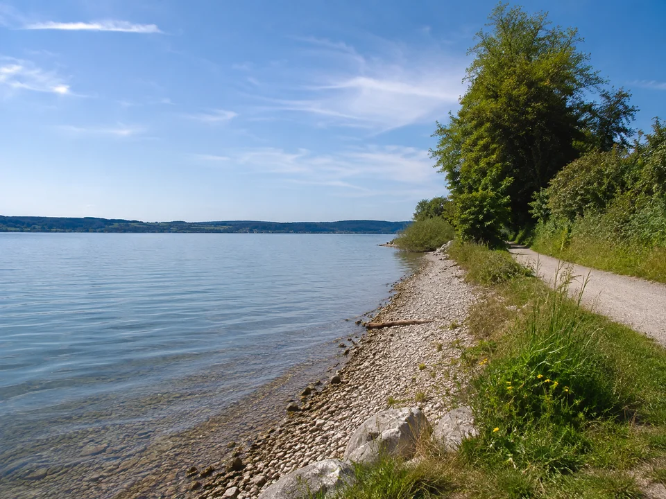 Ammersee bei Utting: natürlicher Kiesrand neben öffentlichem Uferweg, ruhiges Wasser und niedrige gegenüberliegende Moränenhügel