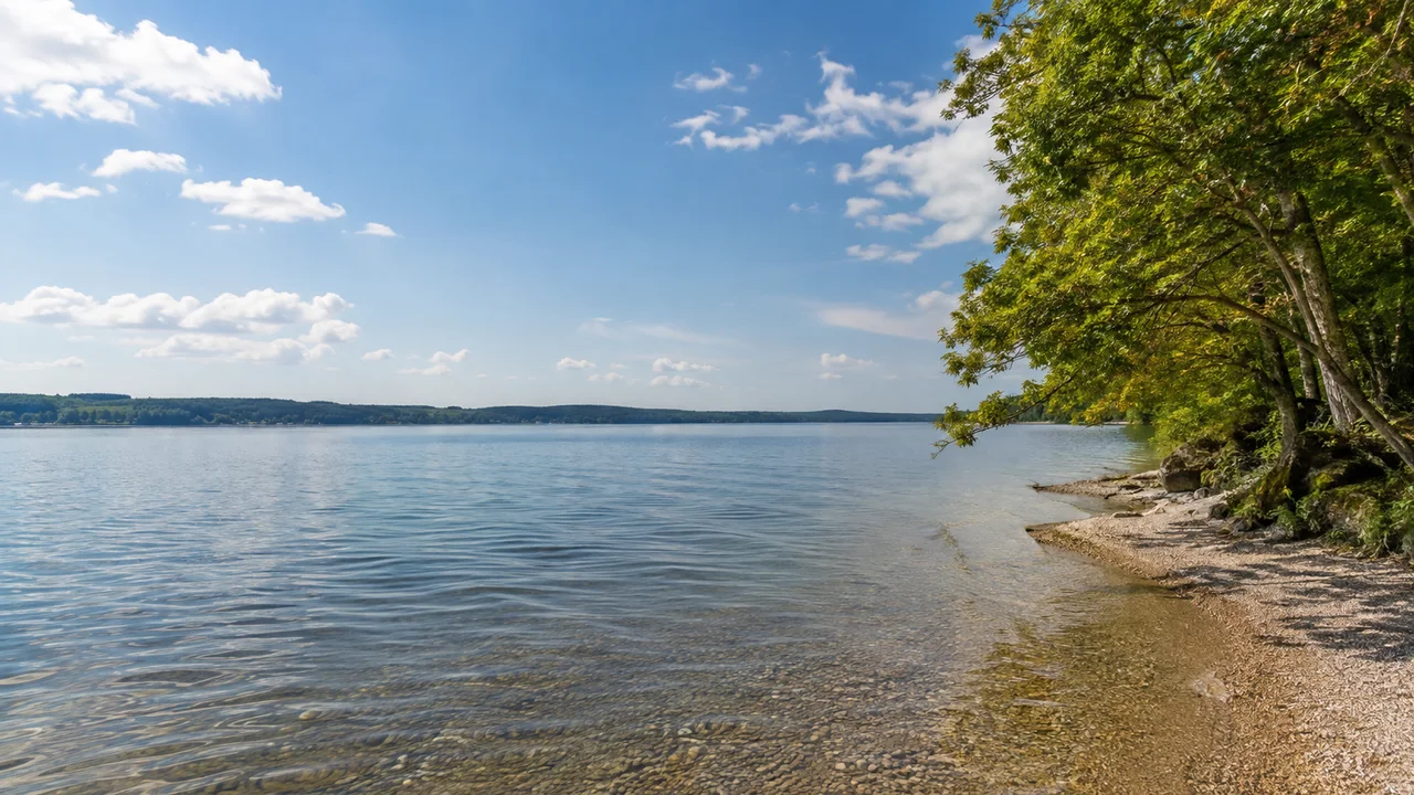 Ammersee: Herrsching Bahnhof: Ammersee von Stegen: breite offene Wasserfläche nach Süden, niedrige bewaldete Ufer, kleine Kiesbucht mit Laubbäumen