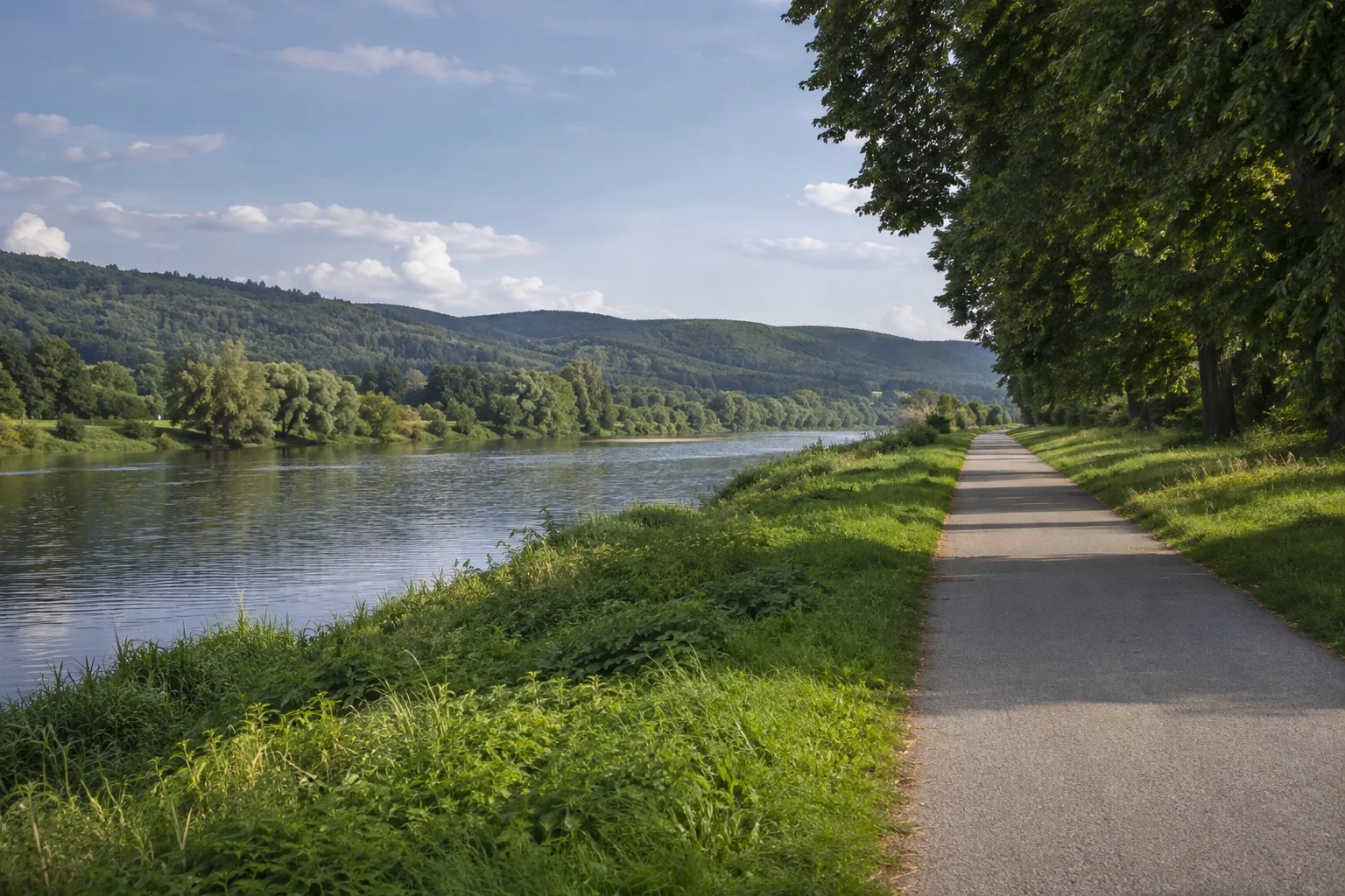 Aschaffenburg: Main bei Aschaffenburg: breiter ruhiger Fluss mit niedrigem grünem Ufer und regulärem Weg, flache Talsohle vor fernen Waldhügeln.