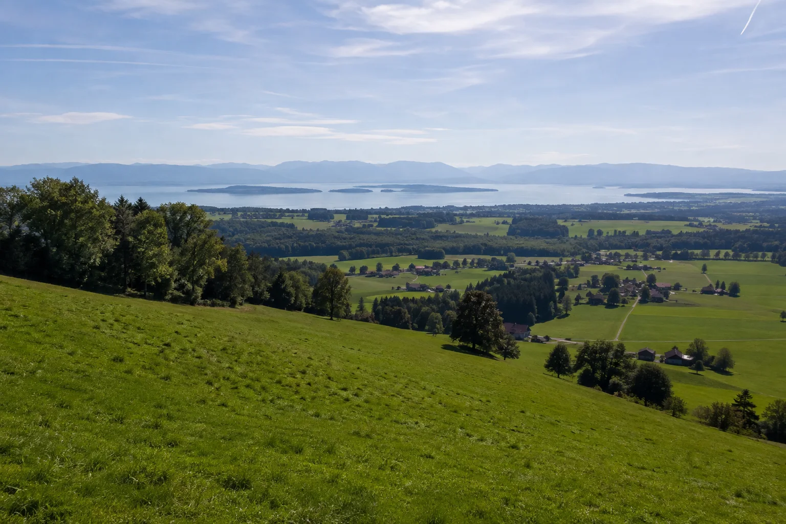 Hittenkirchen: grüner Wiesenhang mit weit unten liegendem Chiemsee und fernen Inselkonturen