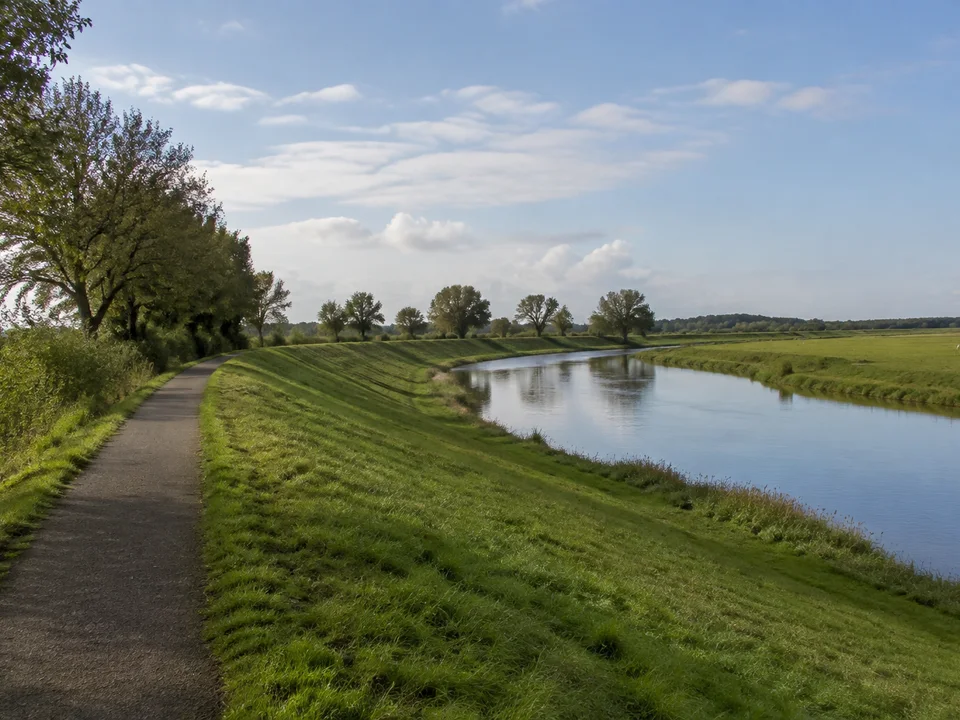 Wümmedeich: Befestigter schmaler Deichweg neben dem windenden ruhigen Wümmefluss, niedrige grüne Wiesen, einzelne Weiden