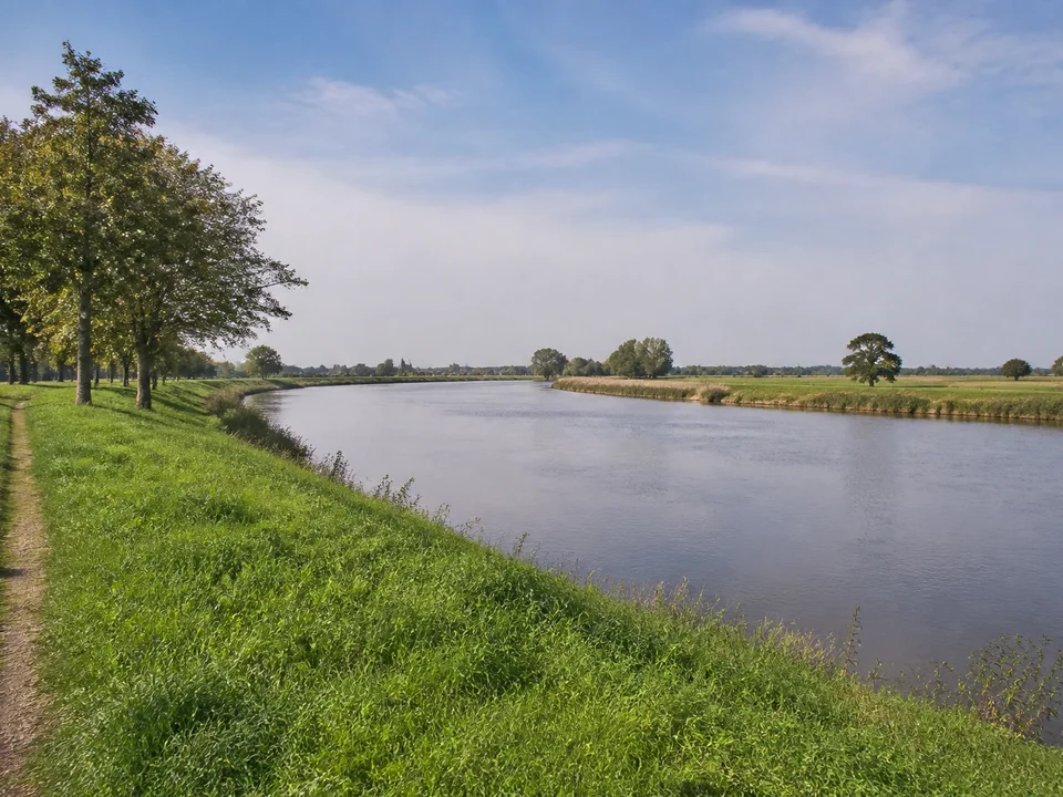 Lesum im Bremer Norden: Breiter ruhiger Gezeitenfluss zwischen niedrigen grünen Deichen, einzelne alte Laubbäume, flache Marschlandschaft