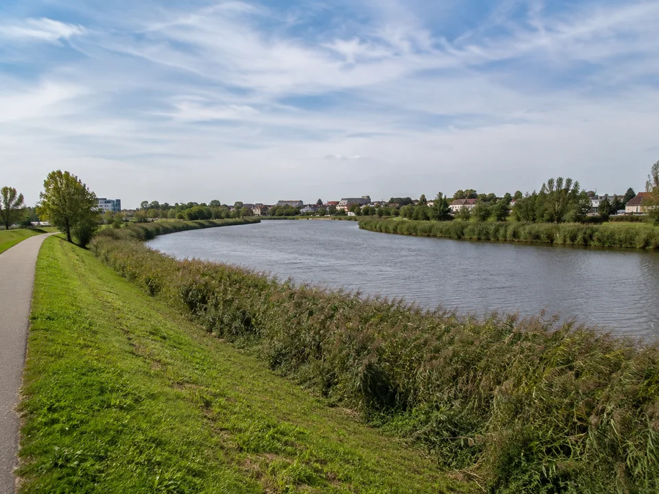 An der Geeste: Ruhige breite Flussschleife mit Schilfrand, Deichweg, grüne Ufer und niedrige städtische Bebauung im Hintergrund
