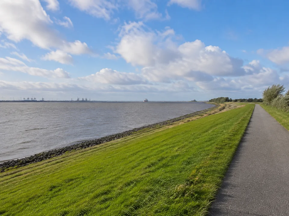 Weser am Seedeich: Breiter graublauer Weserfluss, grasbewachsener niedriger Deich mit befestigtem Spazierweg, ferne Hafenkräne nur klein am Horizont