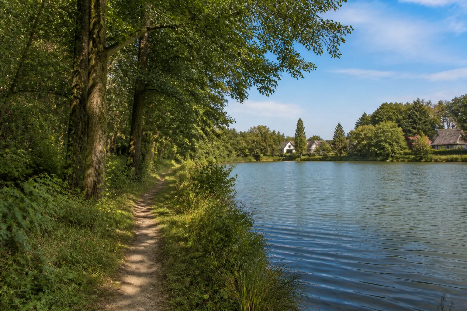 Himmelpforter Seen: schmaler Pfad am bewaldeten Ufer eines kleinen Sees mit einzelnen Dorfhäusern in der Ferne