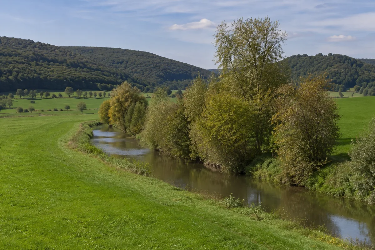 Schmale Leine mit Ufergehölzen zwischen grünen Wiesen westlich Heiligenstadt bei Rengelrode, sanfte bewaldete Höhen
