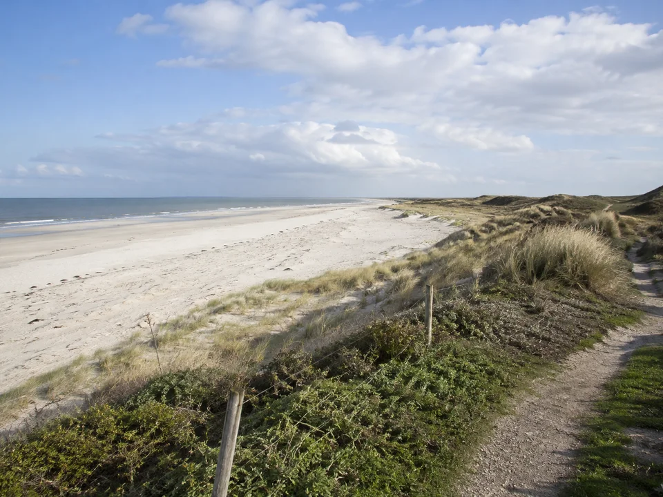 Juist: Breiter heller Nordseestrand am Fuß niedriger Dünen, sanftes Meer und weicher Wolkenhimmel