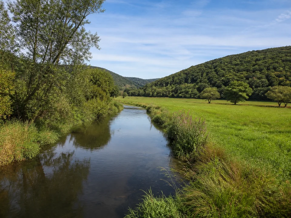Obere Lahnaue: Schmaler ruhiger Fluss zwischen weiten grünen Talwiesen und rundlichen Waldbergen