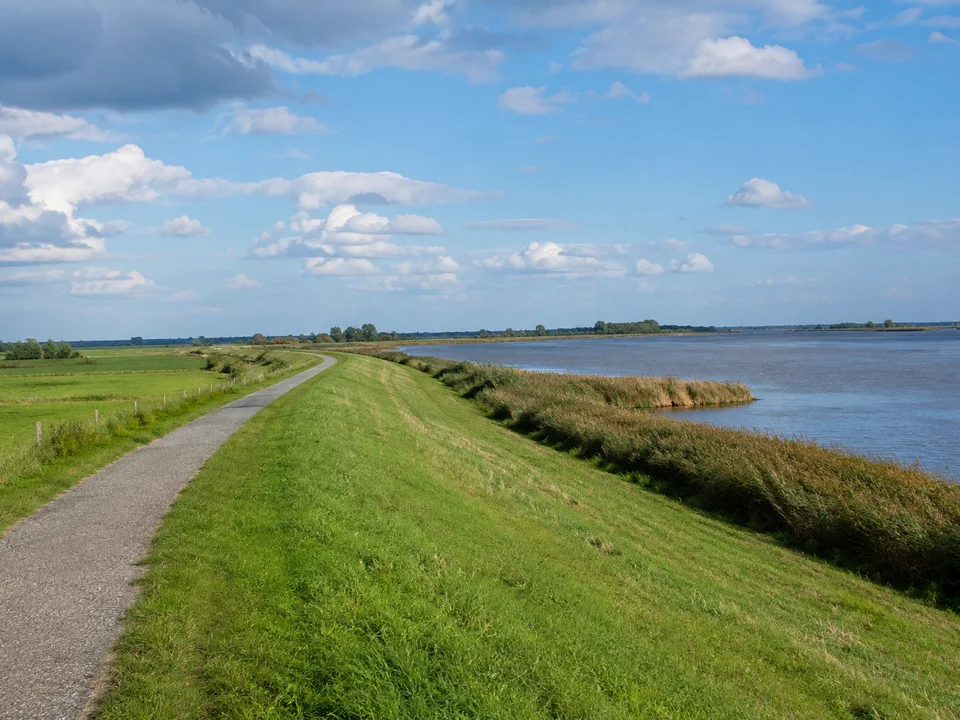 Leer (Ostfriesland): Öffentlicher Deichweg vor einem breiten flachen Fluss, grüne Wiesen und Schilfränder unter weitem Himmel