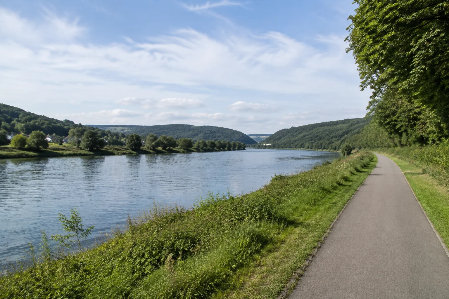 Merzig: Saar bei Merzig: breiter ruhiger Fluss mit niedrigem grünem Ufer, Spazierweg und sanften bewaldeten Hügeln.