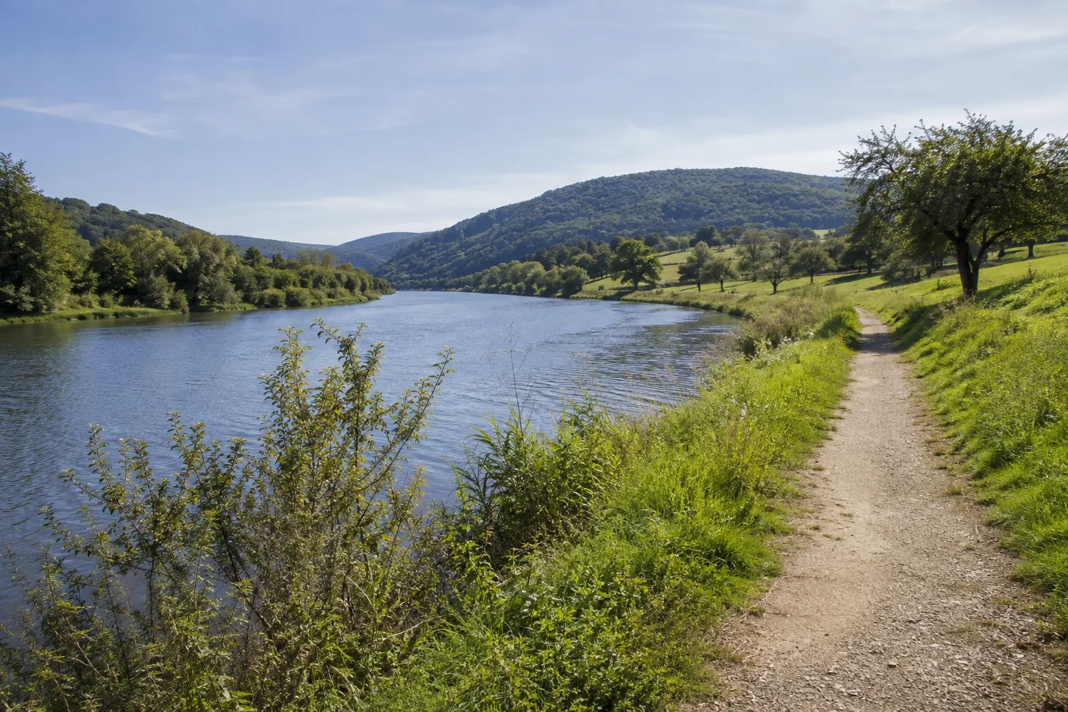 Naturpark Saar-Hunsrück: Merziger Saartal: breiter Fluss in einer offenen Talsohle, Uferweg und Obstwiesen vor niedrigen bewaldeten Hügeln.