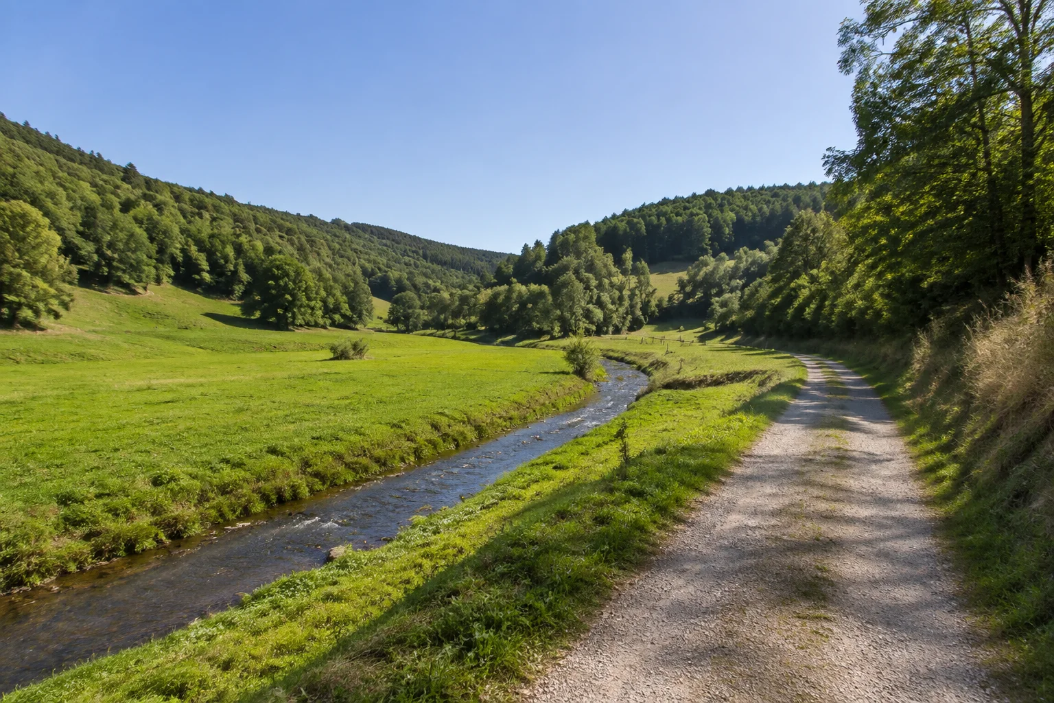 Primstal: kleiner Fluss durch grüne Wiesen zwischen niedrigen bewaldeten Hügeln, Feldweg am Talrand.