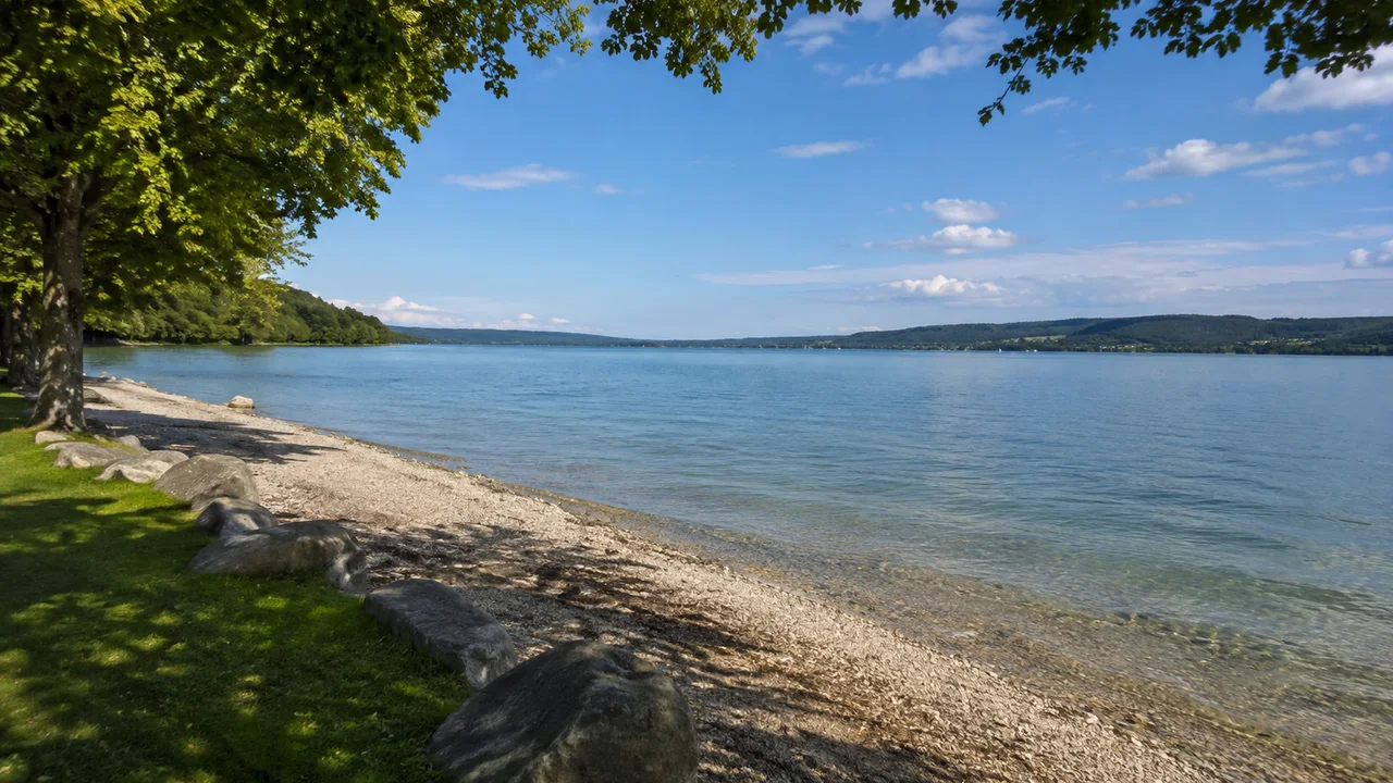 Starnberger See: Starnberg Bahnhof: Starnberger See am nördlichen Ufer: weite Wasserfläche zwischen niedrigen bewaldeten Ufern, heller Uferkies und Parkbäume